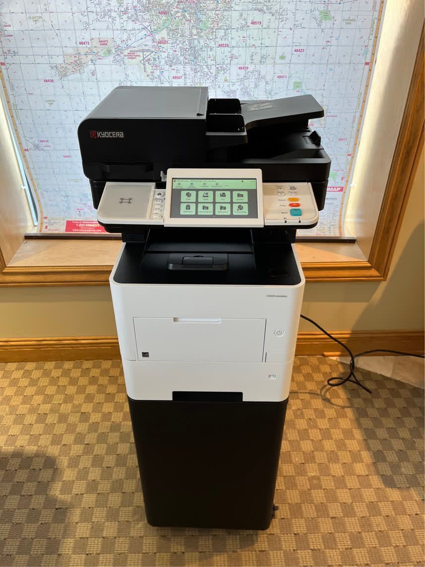 Black and white multifunction printer on a stand, with control panel. Window and map in the background.
