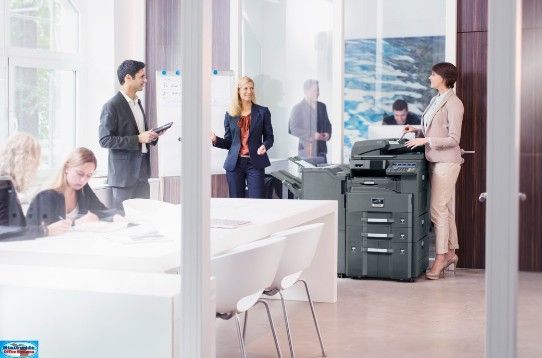 Office setting: people around a copier; woman using it, others in meeting and working.