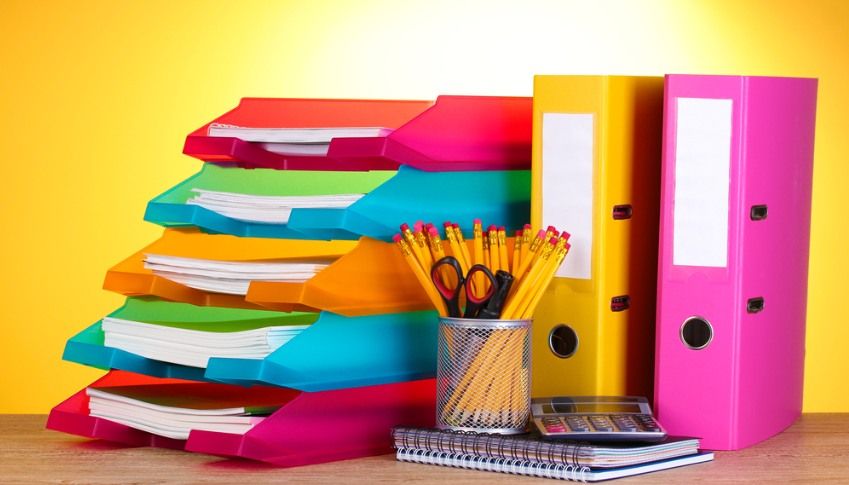 Stack of colorful paper trays and binders, with pencils, scissors, and a calculator on a desk.