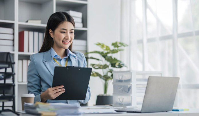 Woman in a blue blazer smiles while holding a clipboard, working on a laptop in an office.