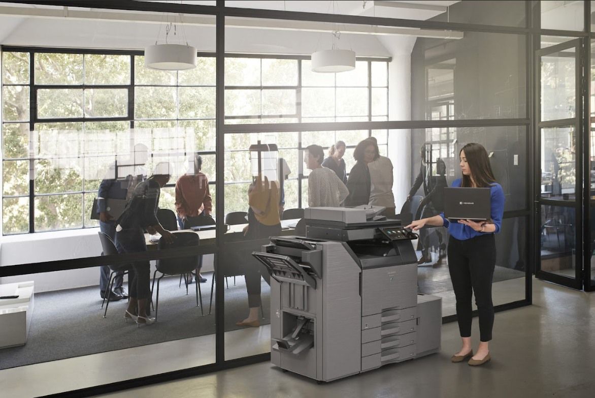 Woman using a printer in an office setting, with a group of people working in the background.