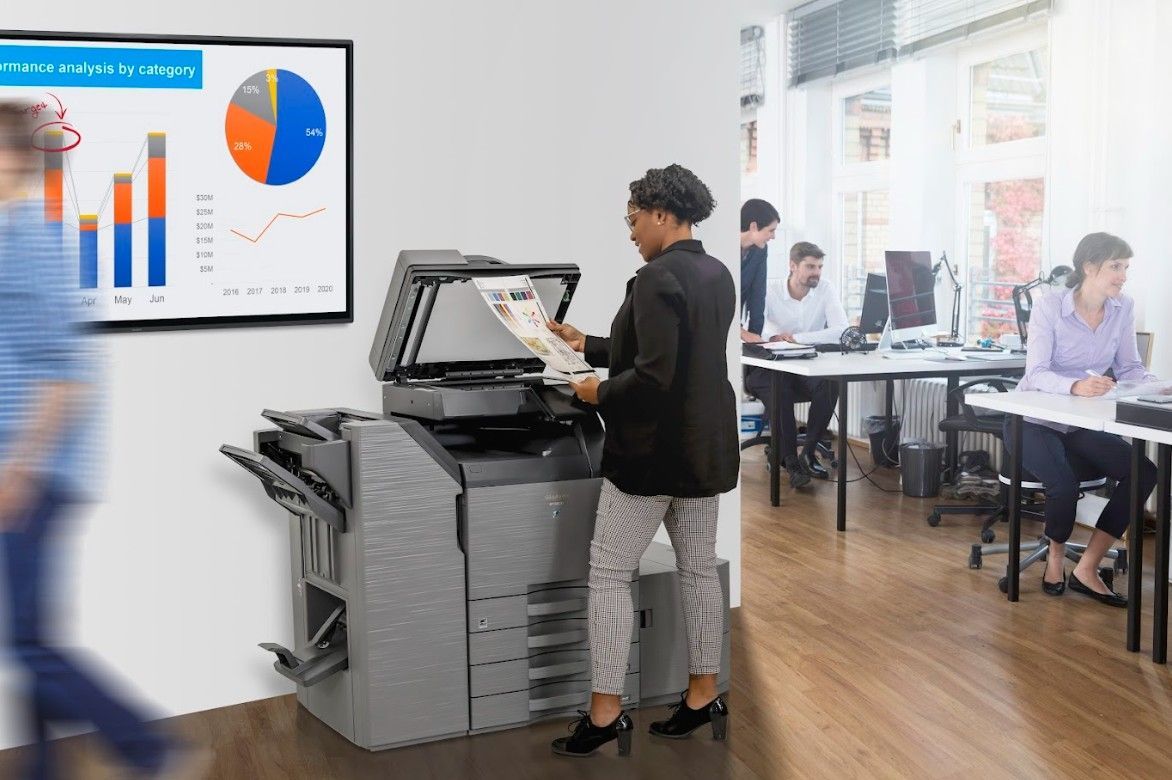 Woman scanning documents on a multifunction printer in an office setting with other employees working on computers.