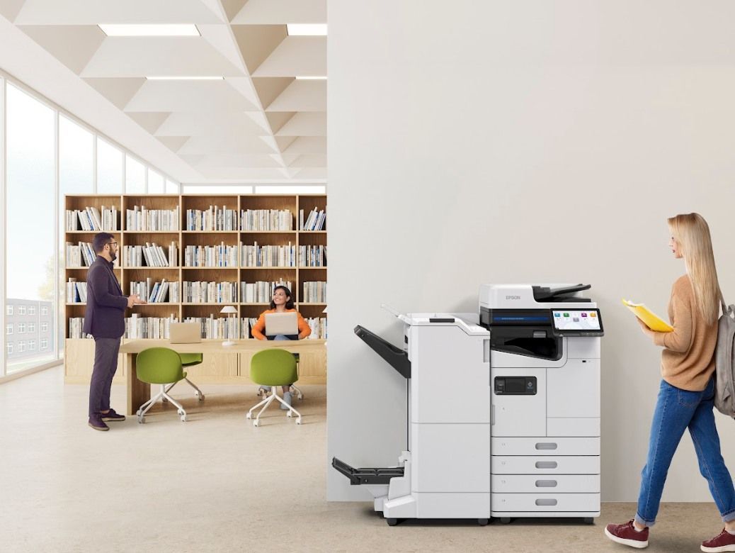 Woman walking by a large office printer in a library setting; a person is working at a computer.