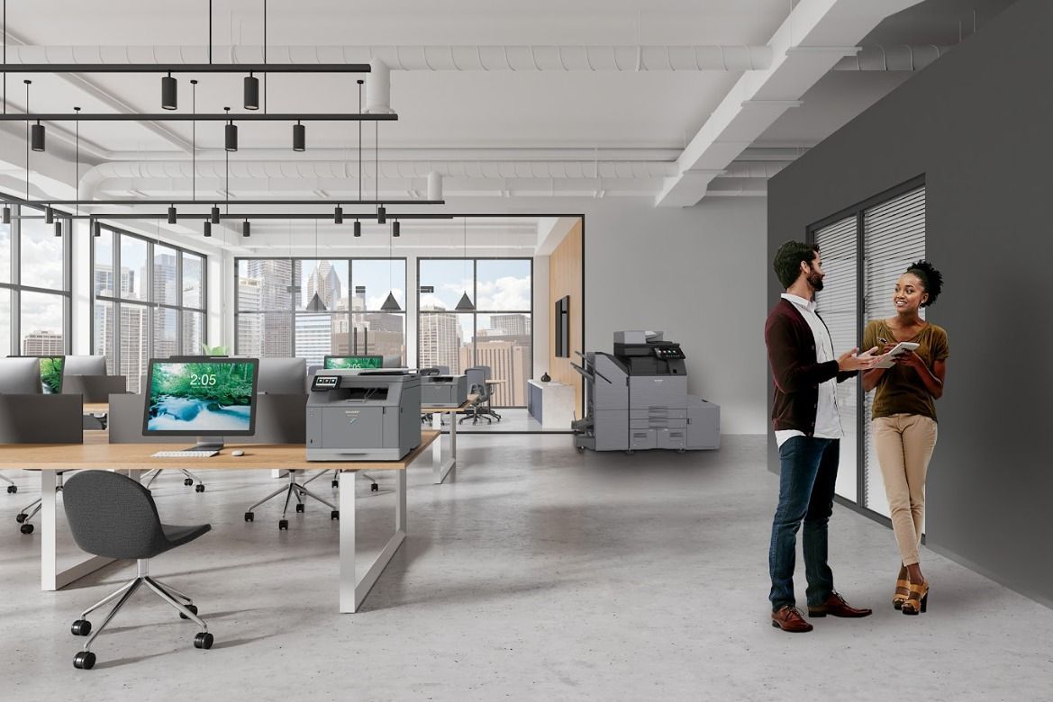 Two people standing near a printer in a modern office with desks, computers, and windows.