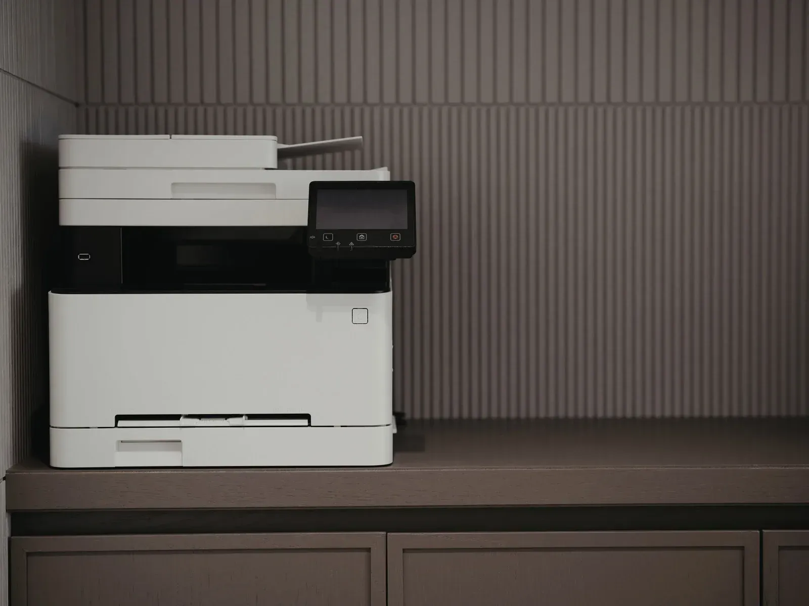 White printer on a dark gray counter in front of a gray ribbed wall.