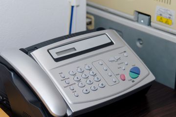 Silver fax machine with a phone handset on a desk, next to a printer.