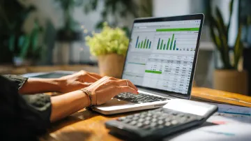 Woman working on a laptop, examining financial data. A calculator sits on the desk next to the computer.