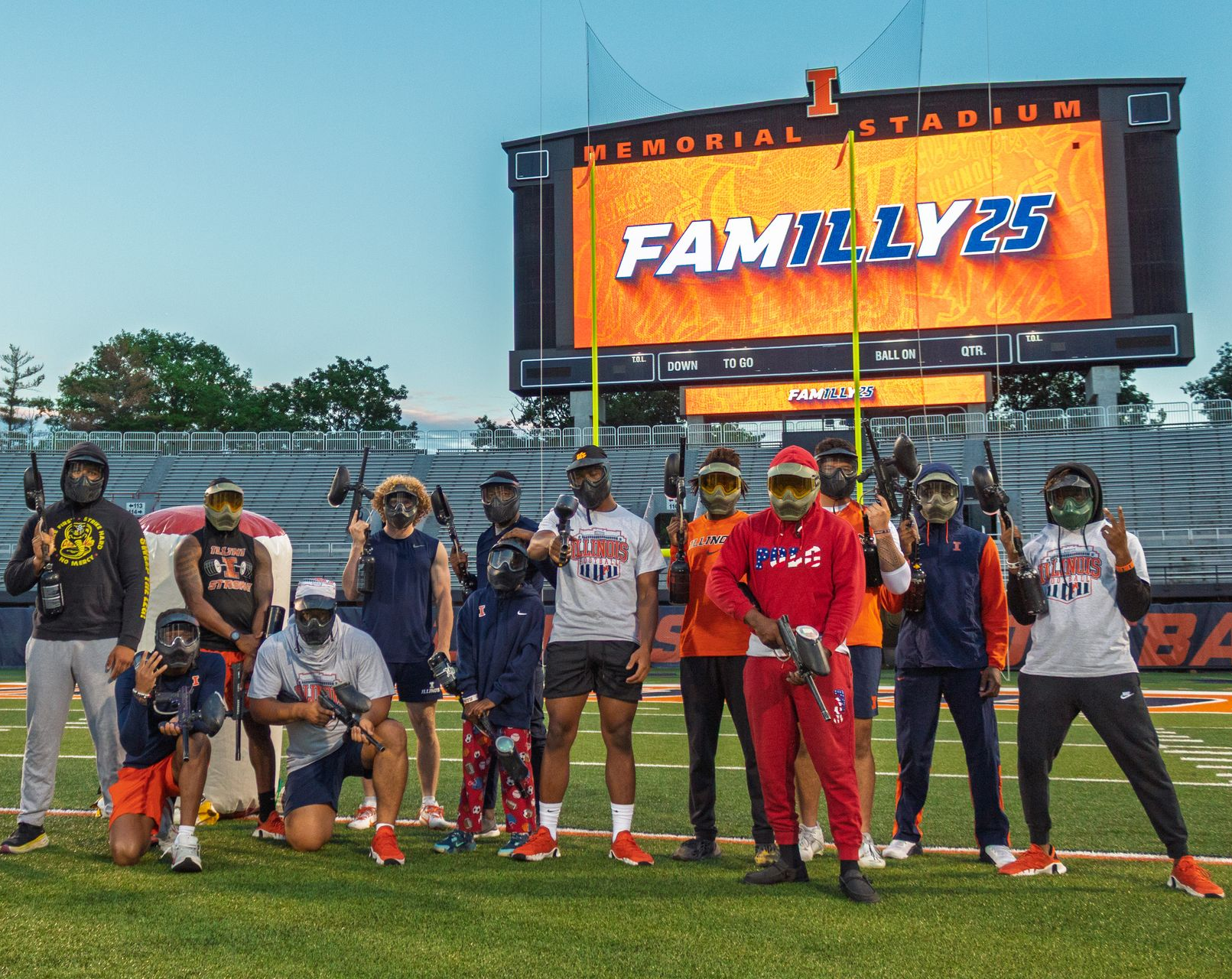 Group of people with paintball guns at a stadium, in front of a scoreboard that reads FAMILY 25.