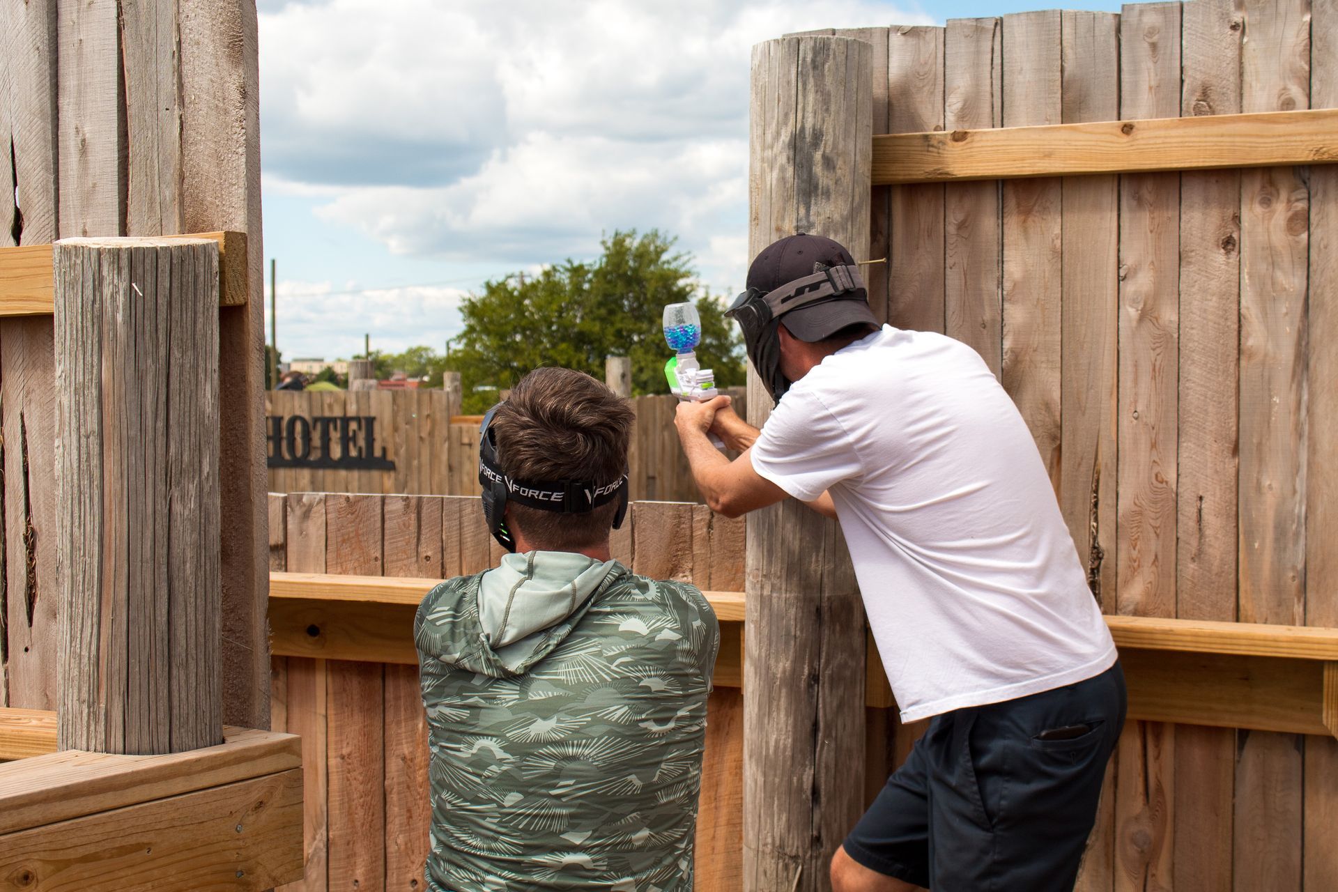 Two people playing paintball behind a wooden barrier; one aiming, the other looking.