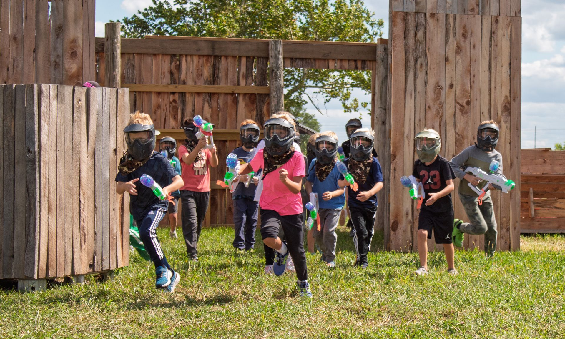 Children wearing protective gear run during a paintball game. Wooden structures, grassy field.