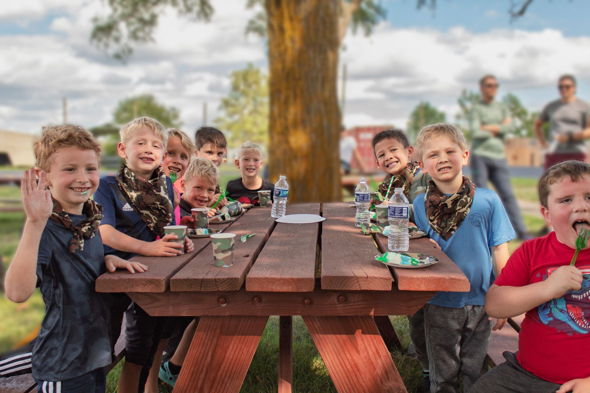 Children at a picnic table, some wearing camouflage scarves. A boy in red eats a snack. Outdoors.