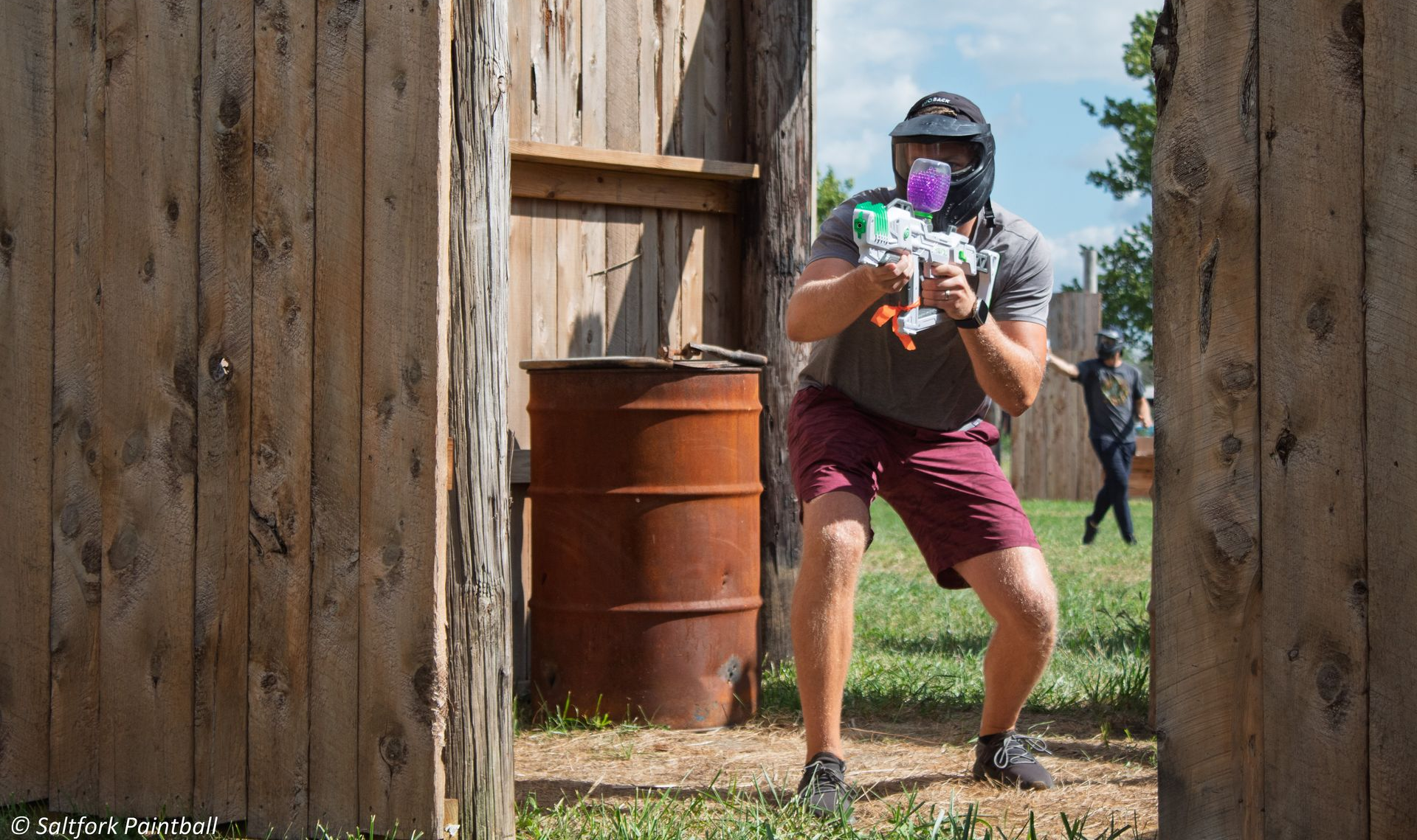 Man in paintball gear aiming a paintball gun from behind a wooden structure.