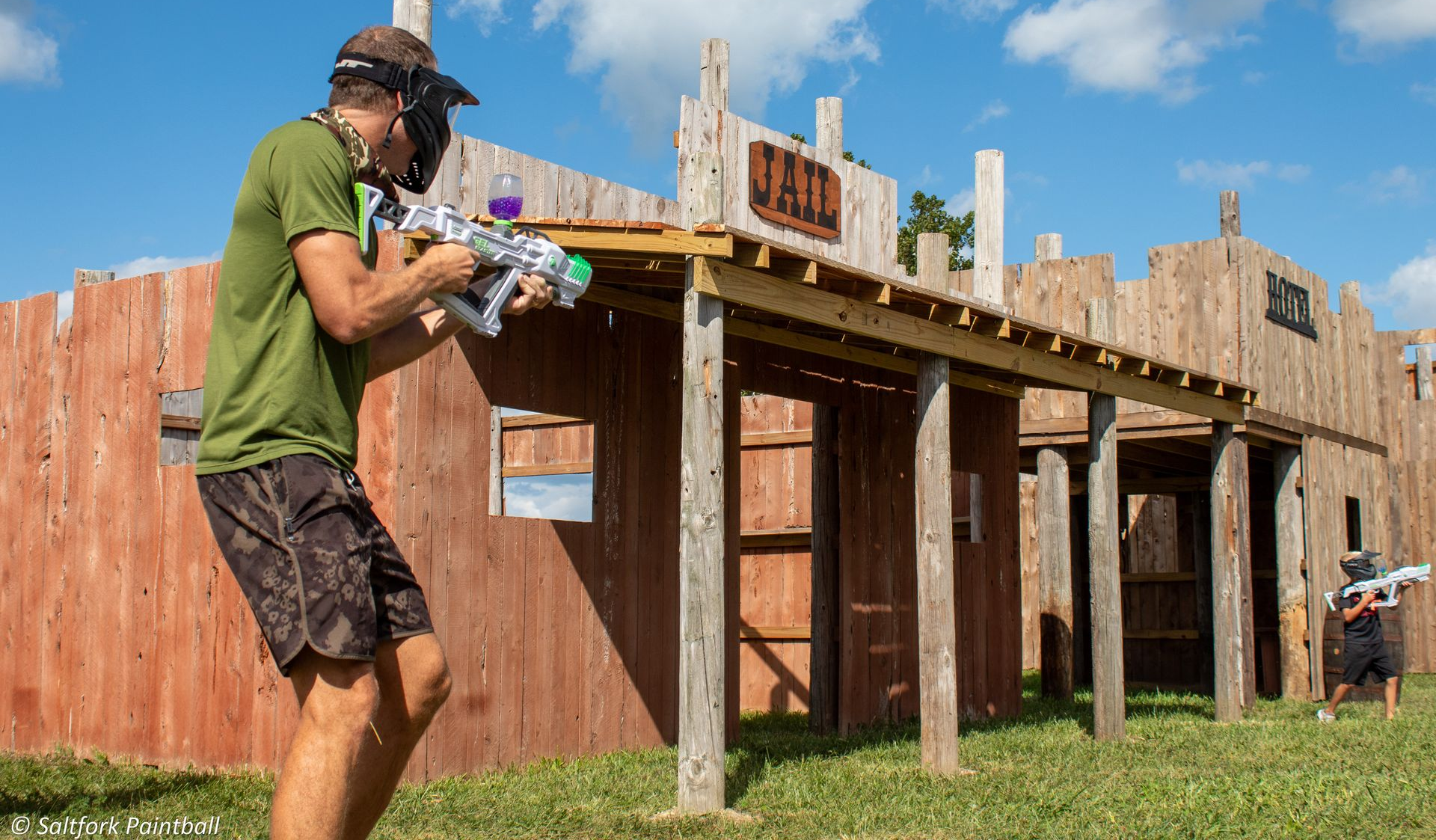 Man in paintball gear aims gun at opposing player in a western-themed arena.