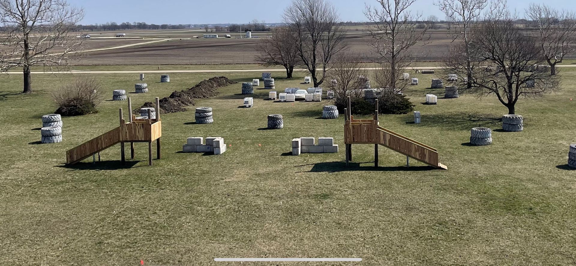 A paintball field with obstacles, ramps, and trees in an open grassy area under a sunny sky.