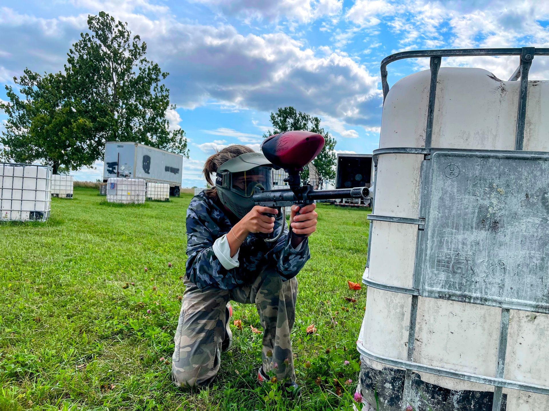 Person in camo gear aiming a paintball gun behind a white barrier on a grassy field under a cloudy sky.