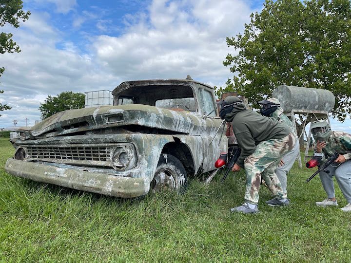 People playing paintball behind a dilapidated truck on a grassy field.