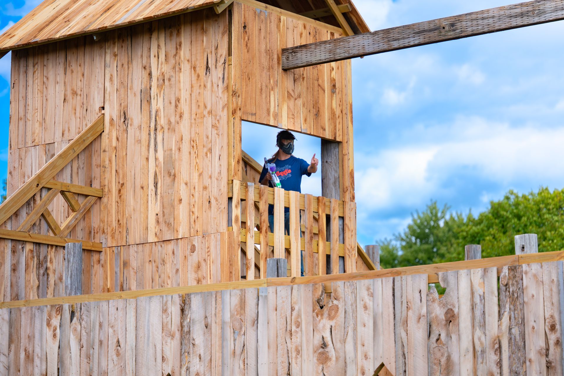 Man with paintball gun in wooden tower, blue sky.