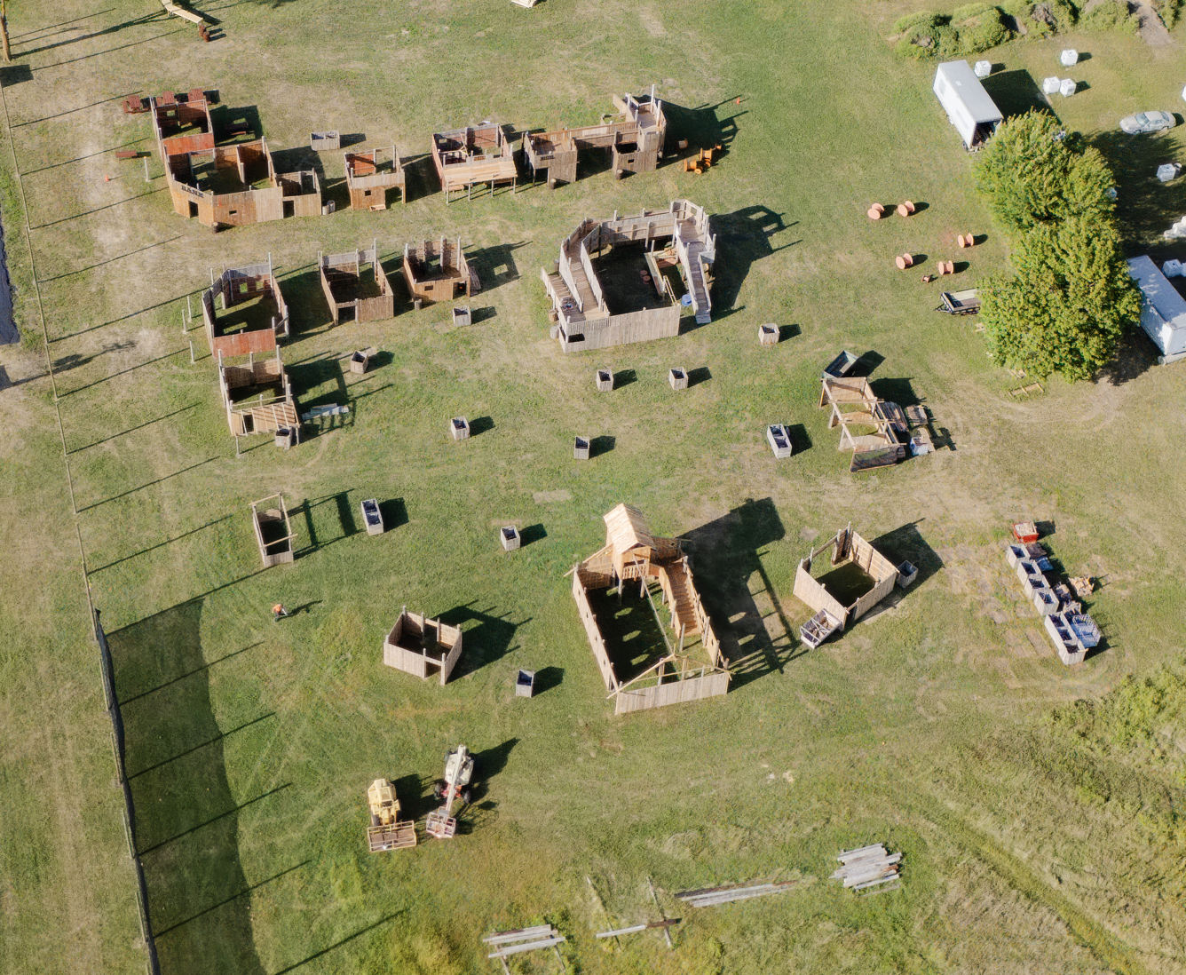 Aerial view of a paintball field with wooden structures on a grassy field; some trees and a trailer are present.