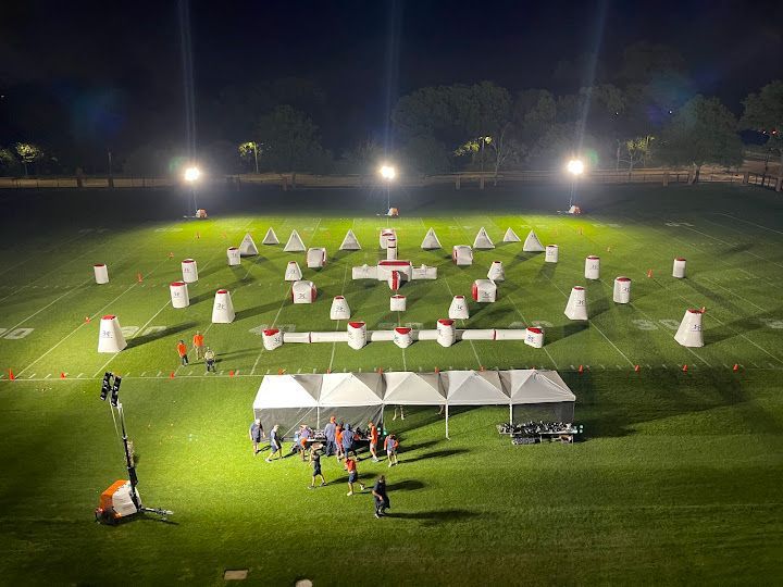 Nighttime paintball field lit by spotlights, with bunkers, tents, and players.