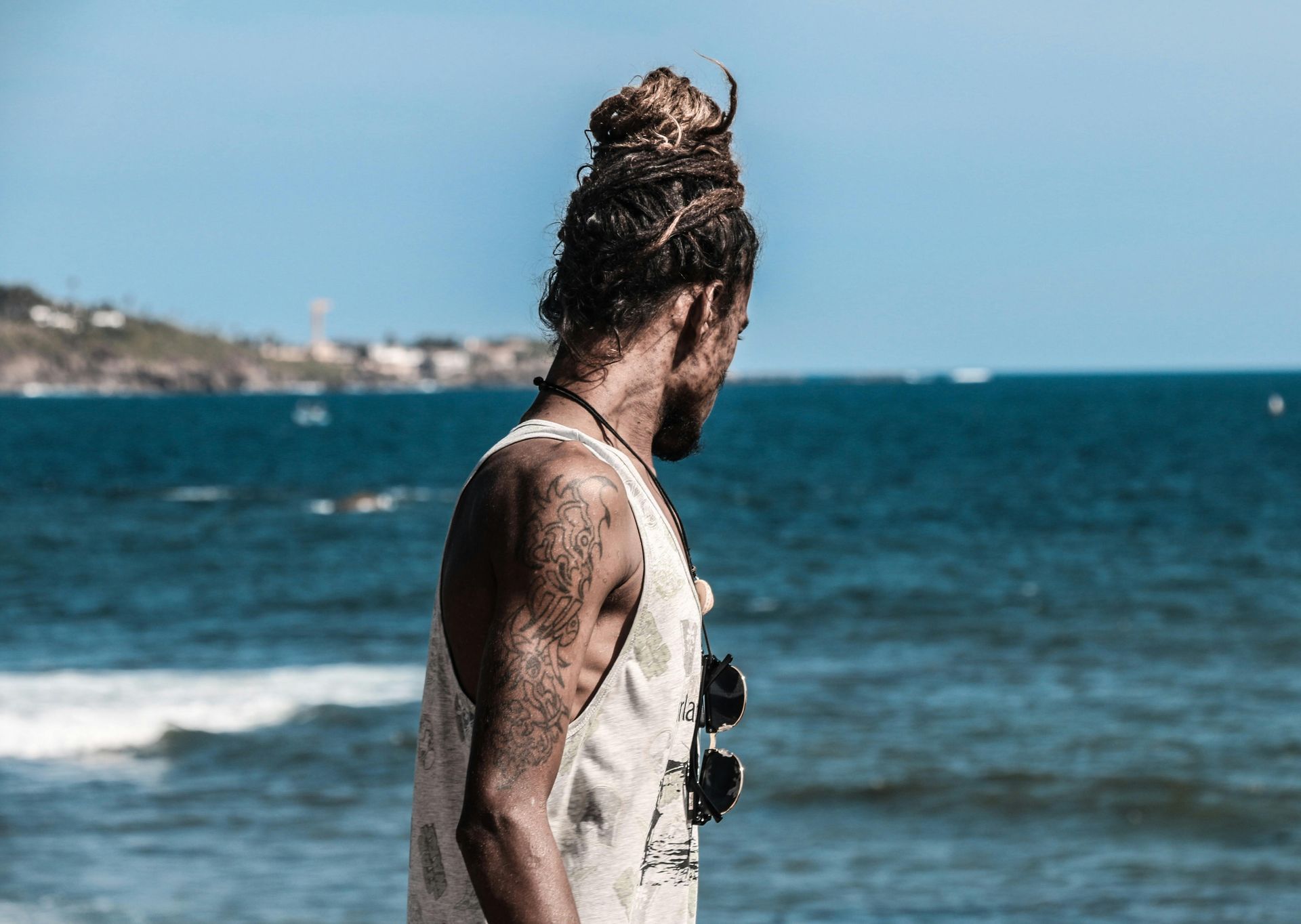 A man with tattoos is standing on a beach looking at the ocean.