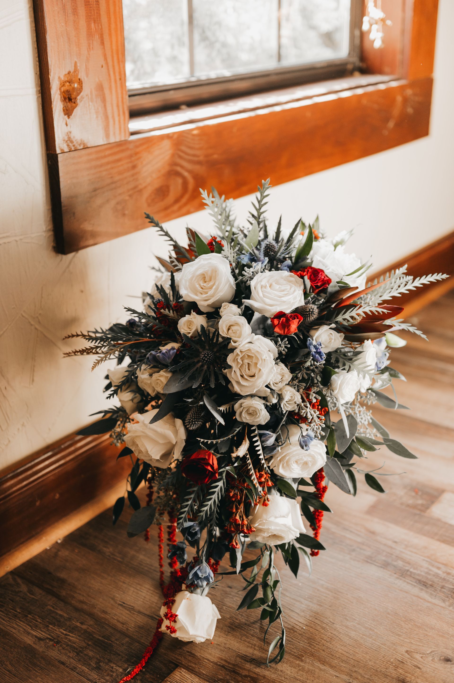 Bridal bouquet with white roses, red berries, blue accents, and greenery near a window.