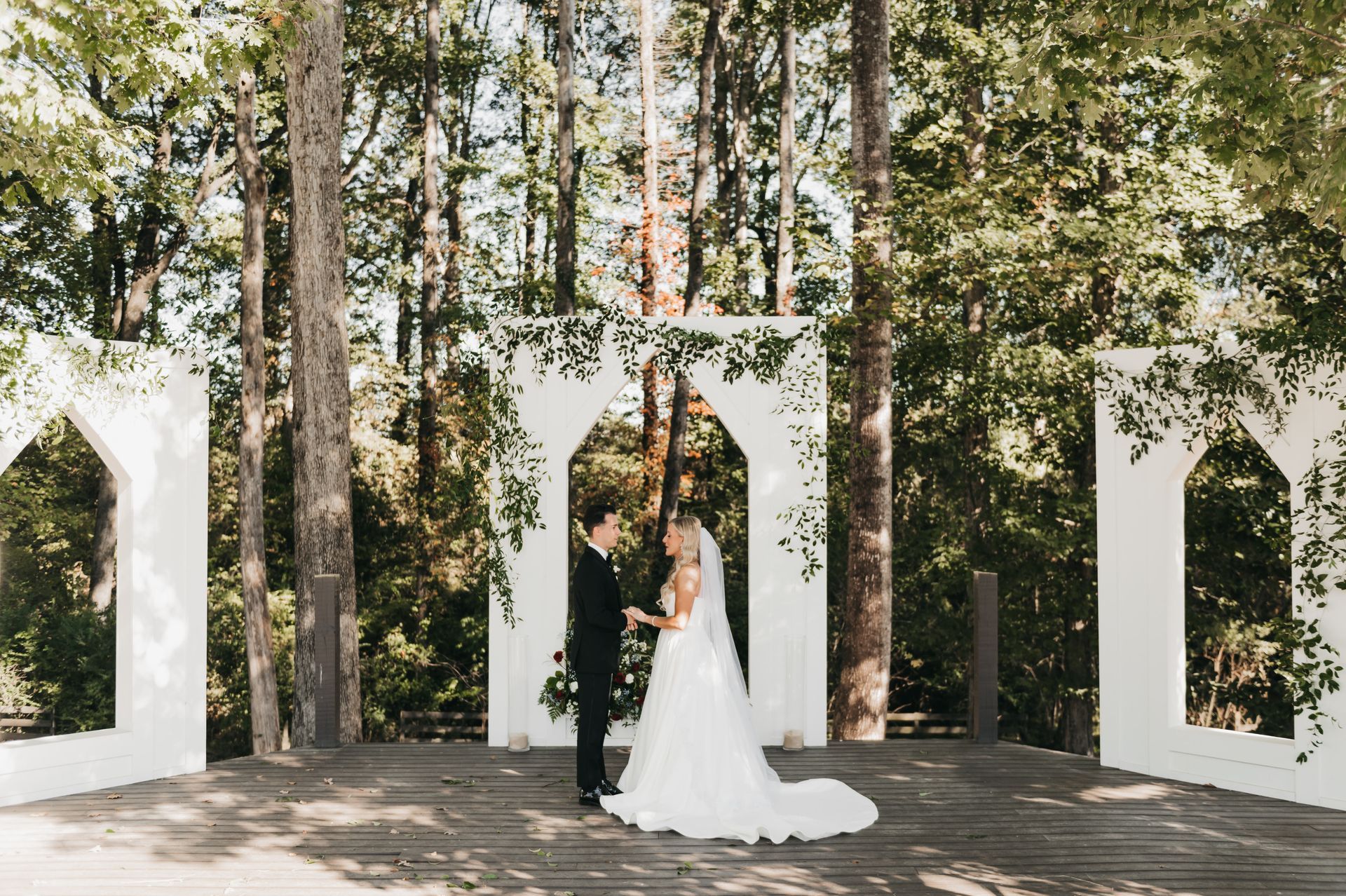 Couple at wedding ceremony, standing before arch, surrounded by trees.