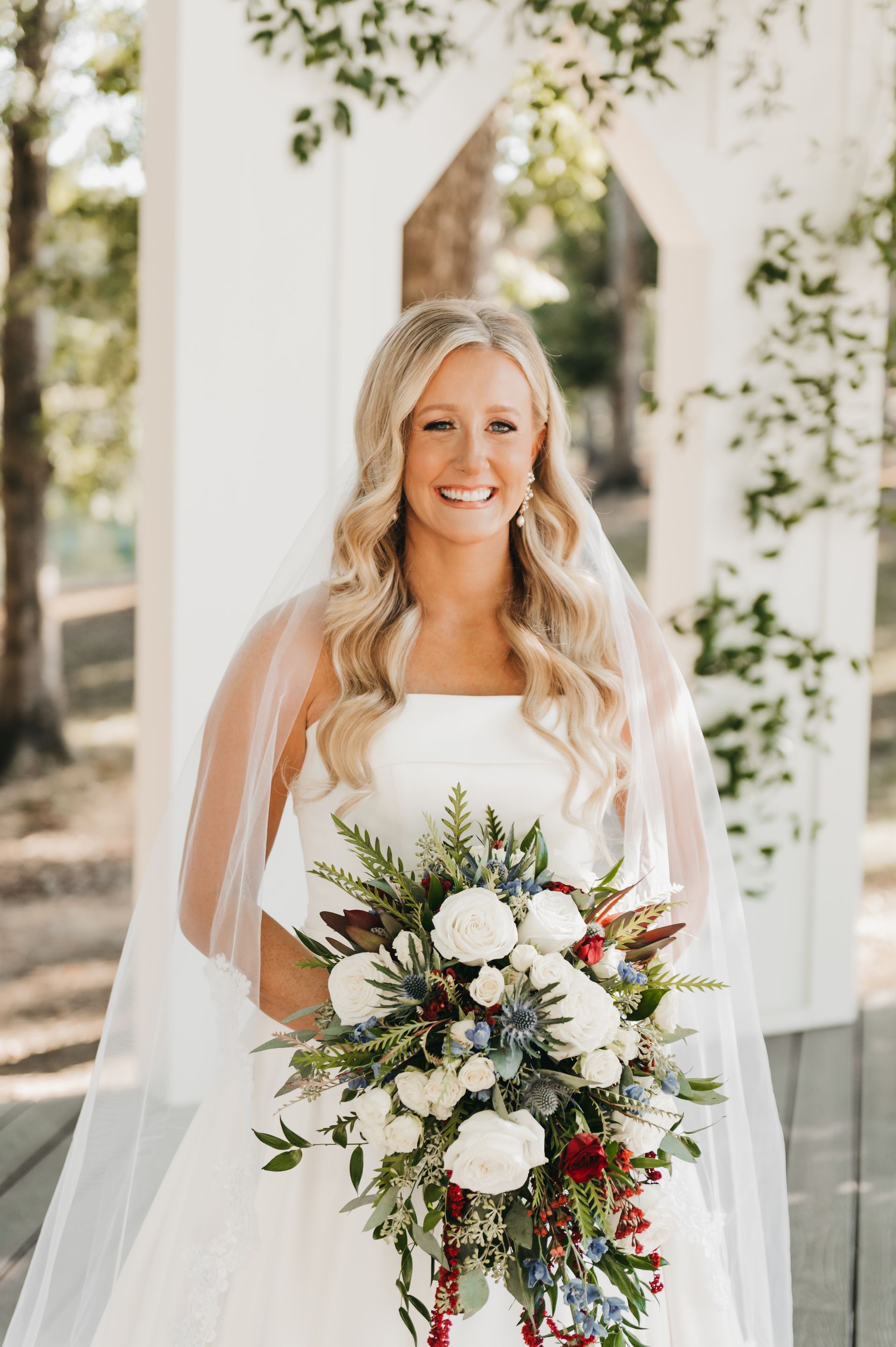 Bride in white dress holding bouquet, smiling, under floral arch.