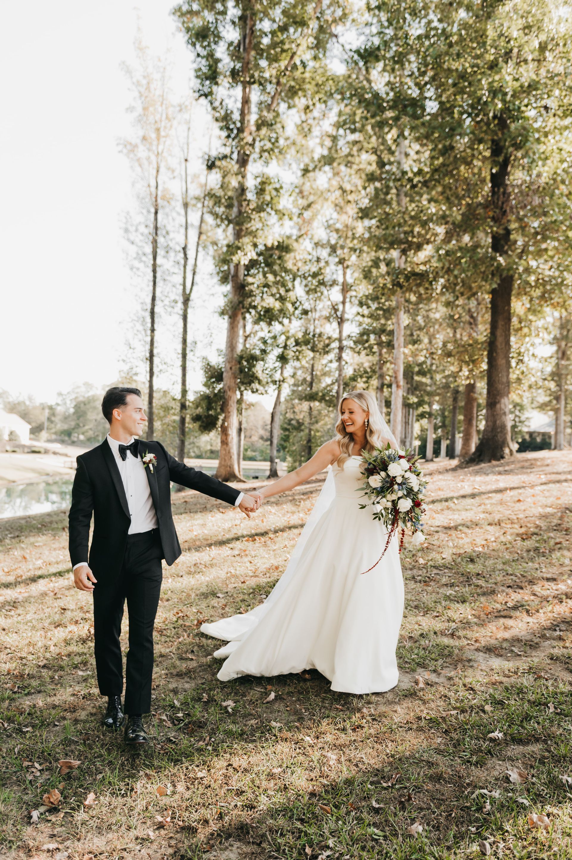 Newlyweds walk hand-in-hand in a sunny field. The bride wears a white gown, the groom a black suit.