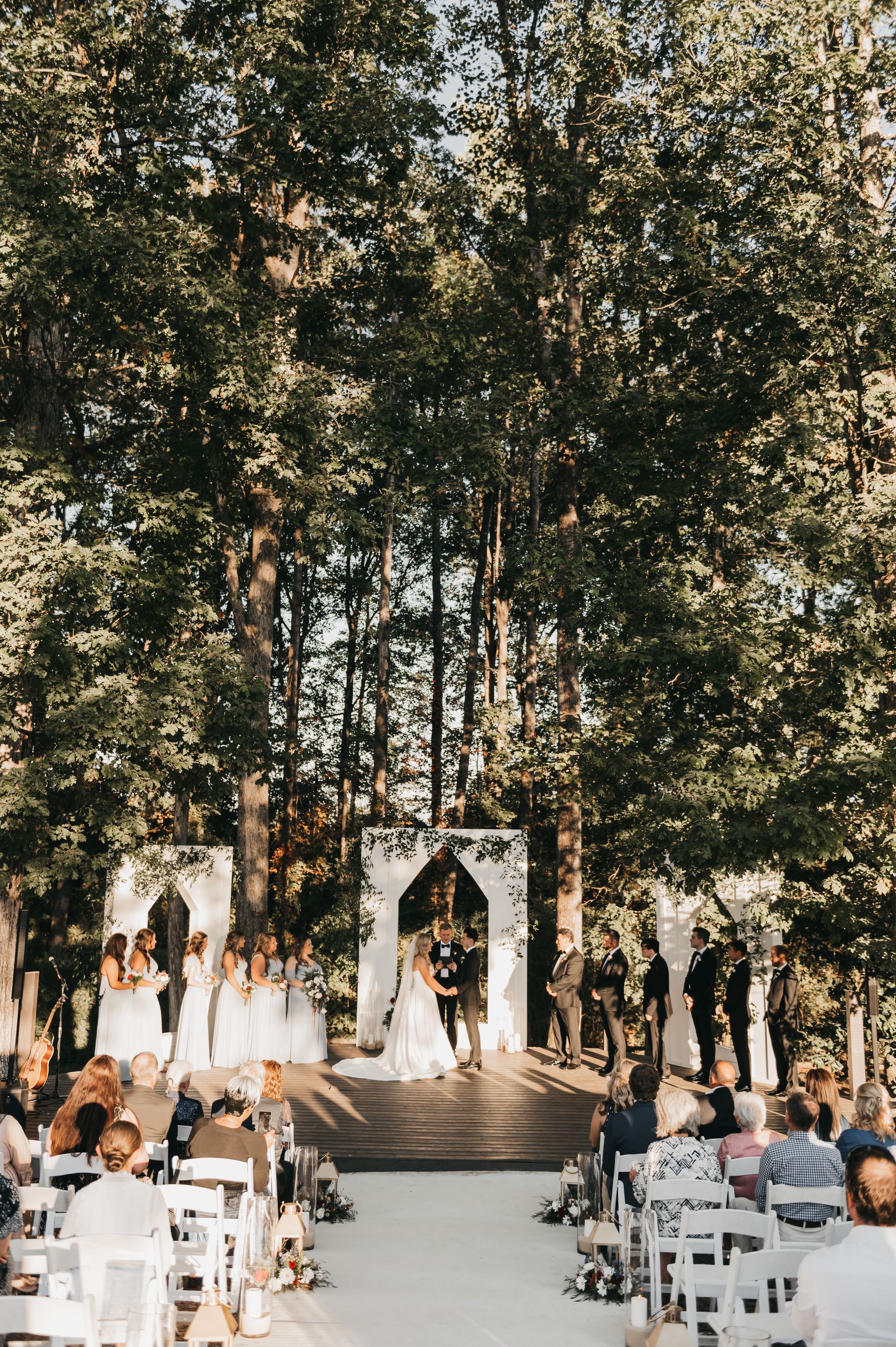 Wedding ceremony outdoors, couple at altar, surrounded by wedding party and guests. White archway and lush trees.
