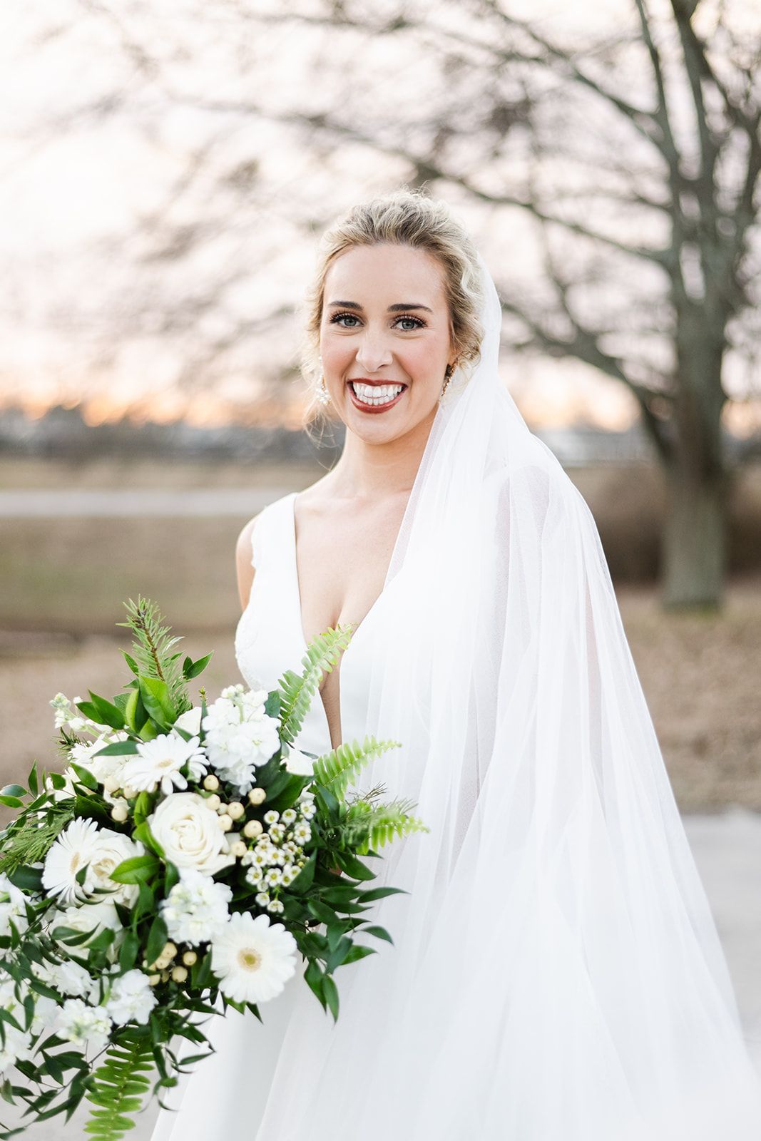 Bride smiling, holding white bouquet, wearing a white wedding dress and veil outdoors.