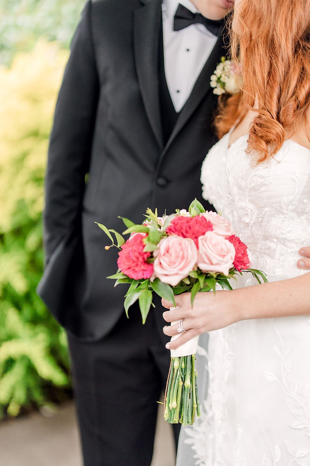Bride holding bouquet, in white dress, groom in black tuxedo, outdoor greenery in background.