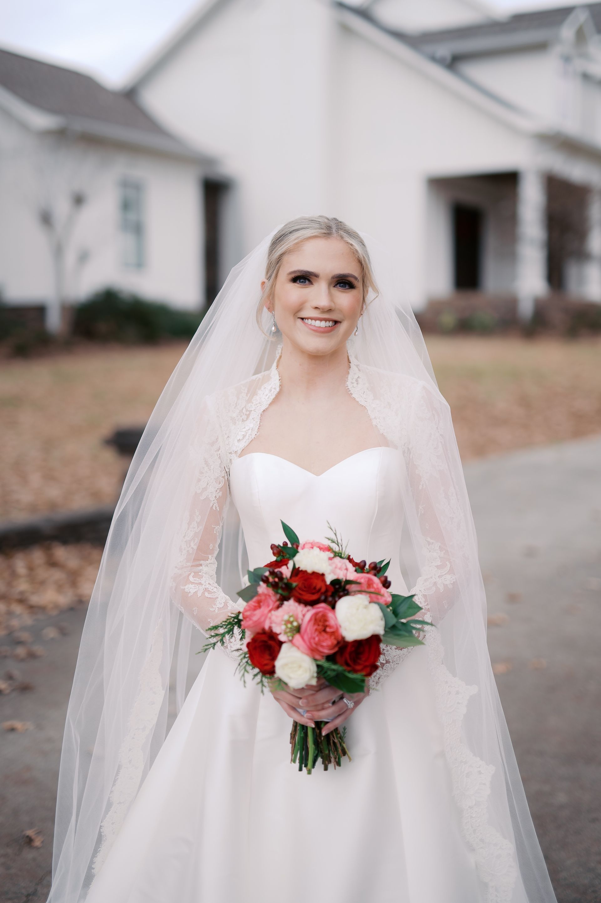 Bride in white dress holding bouquet, smiles outdoors. White veil and building in background.
