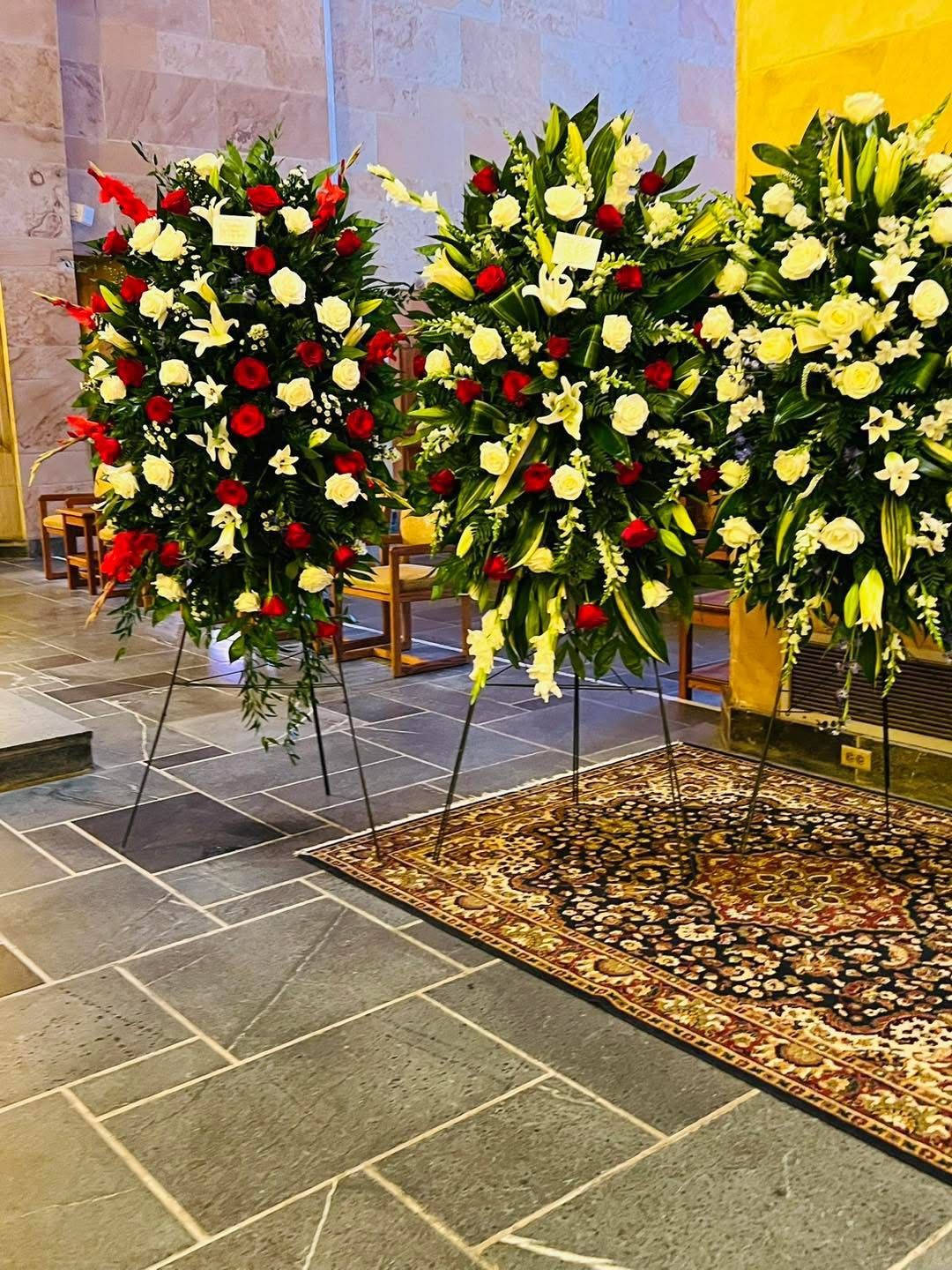 Three large floral arrangements on stands in a building. Red roses and white lilies are prominent, on a stone floor.