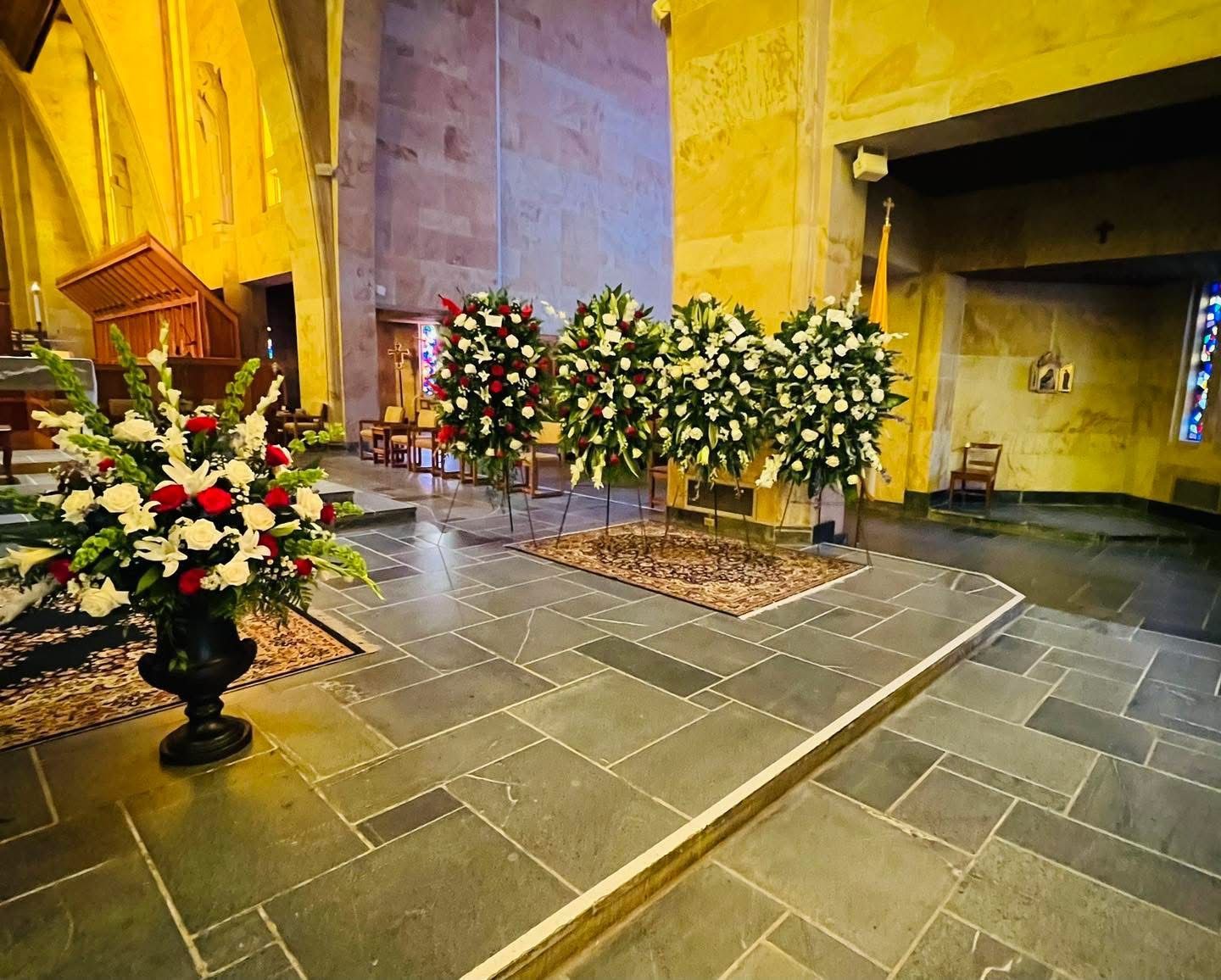 Interior of a church with floral arrangements and stone floor. Three wreaths and a bouquet rest by a grave.