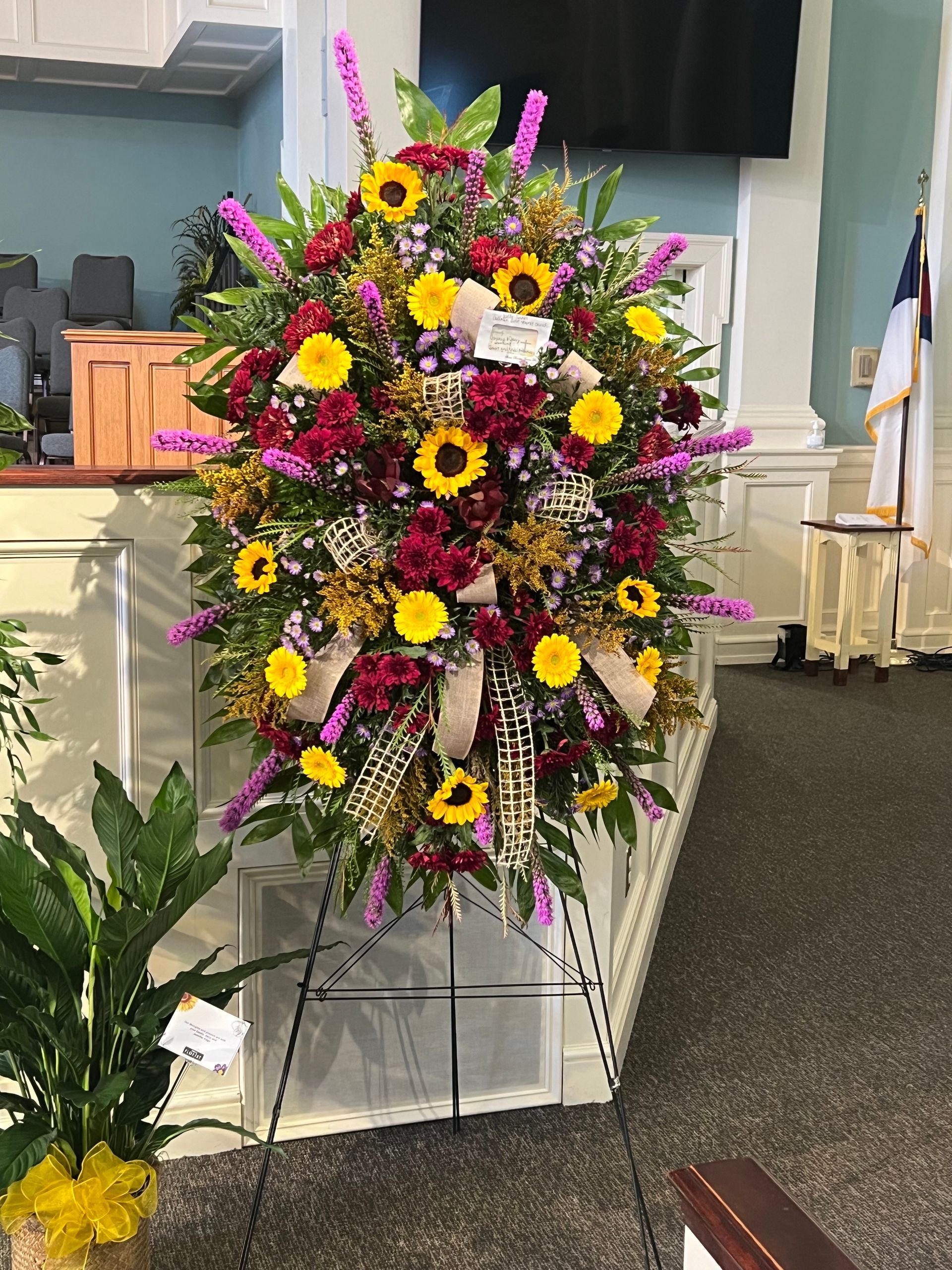Floral arrangement on a stand in a church, featuring sunflowers, burgundy, and purple blooms.