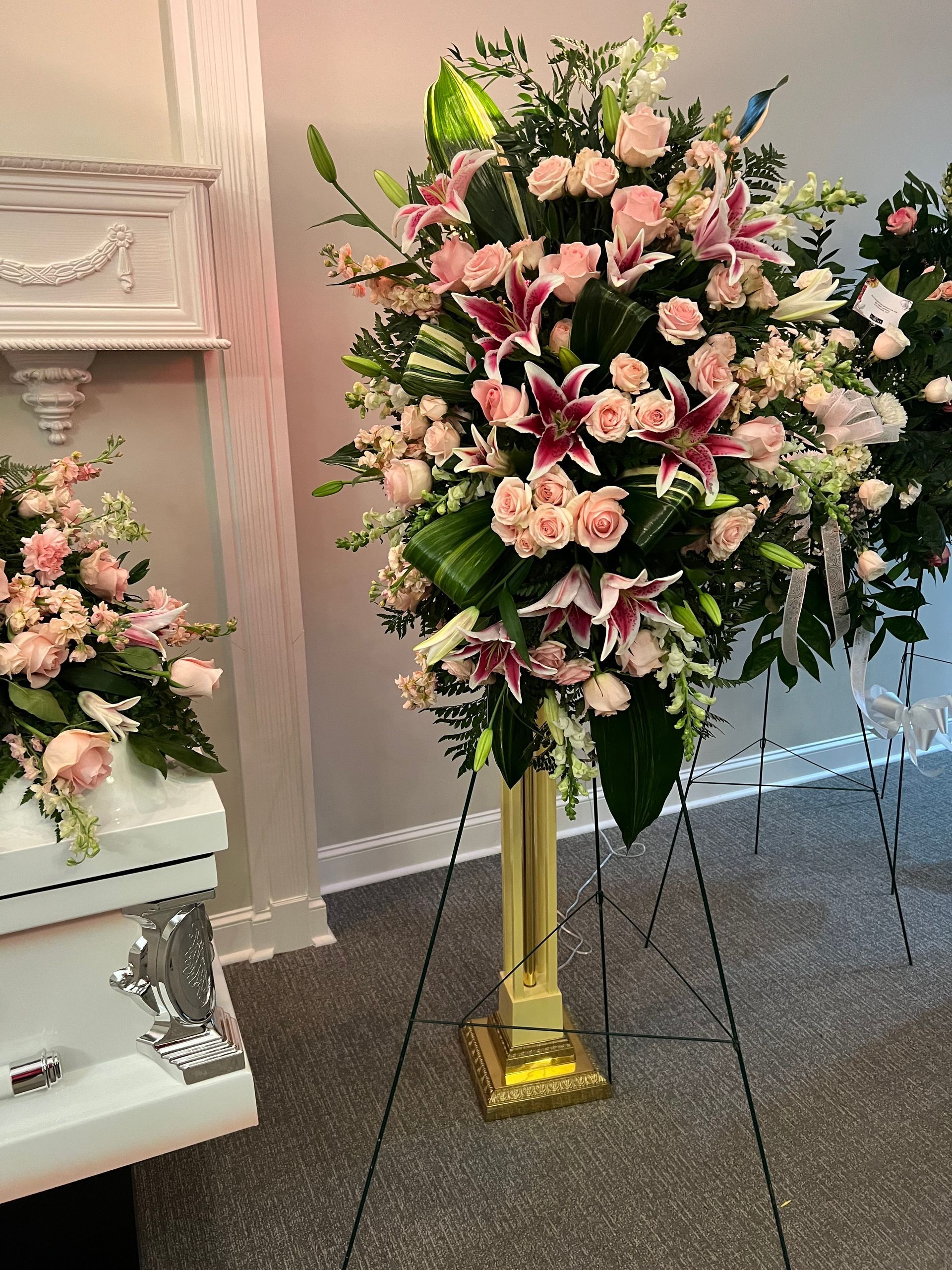 Funeral flower arrangement, pink lilies and roses on a stand, next to a white casket.