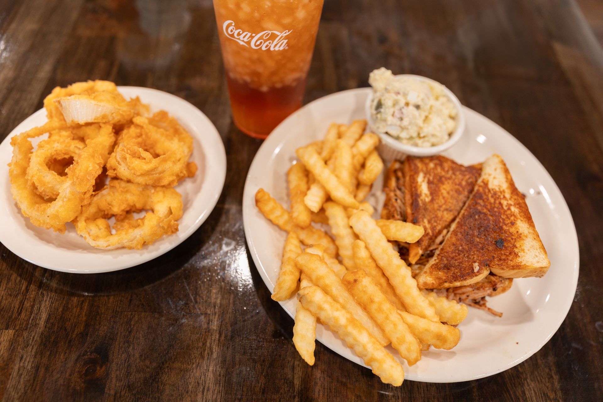 Plates of fried onion rings, fries, and a sandwich with coleslaw; Coca-Cola drink.