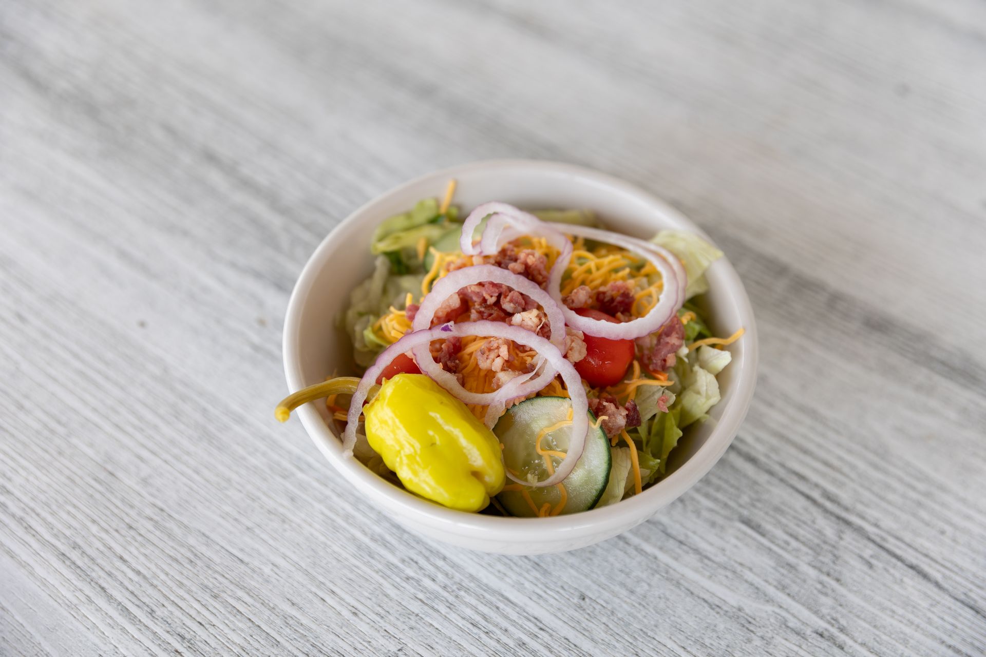 Salad in a white bowl, with lettuce, onion rings, tomatoes, and pepperoncini.