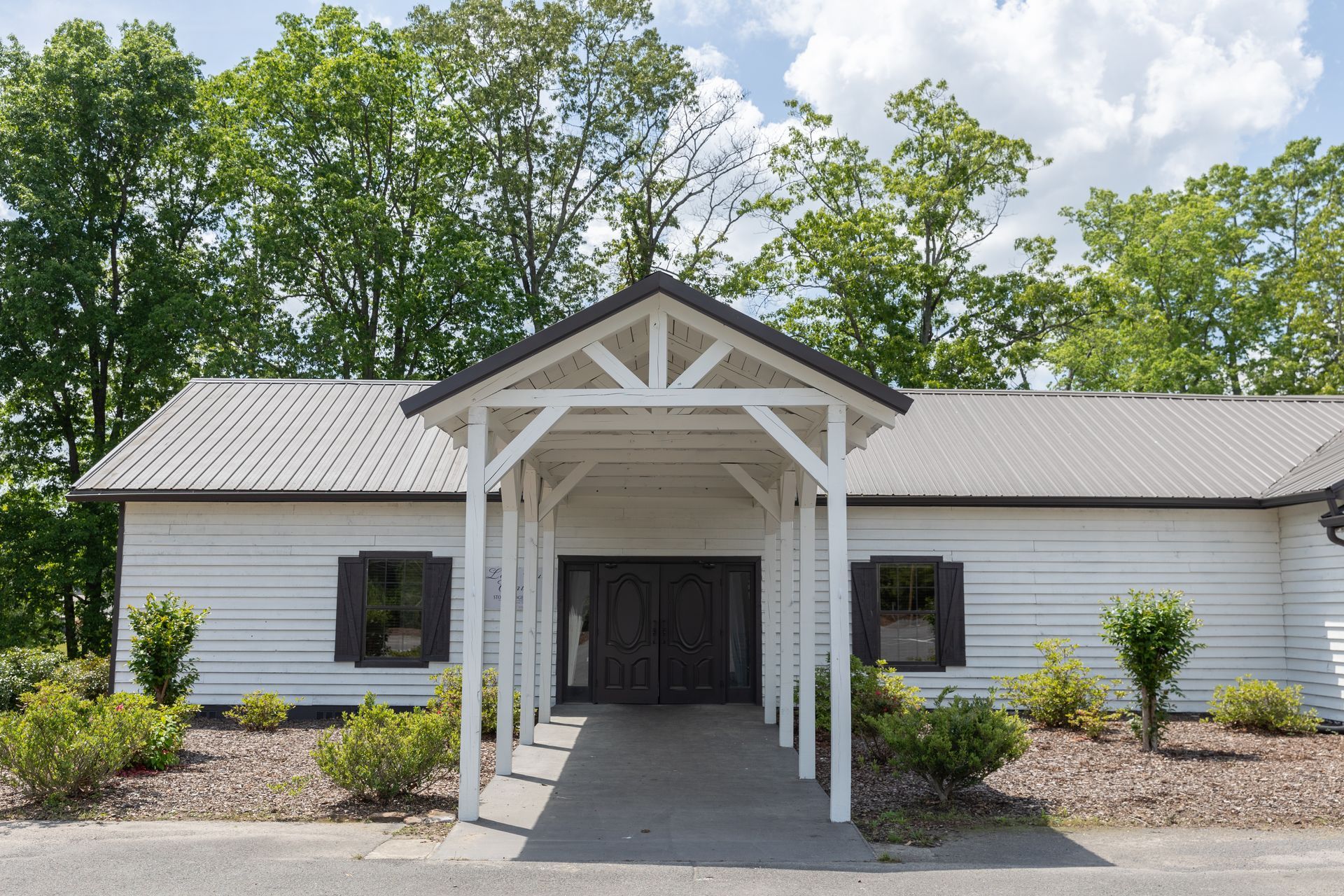 White building with a covered entrance, set against trees and a blue sky.