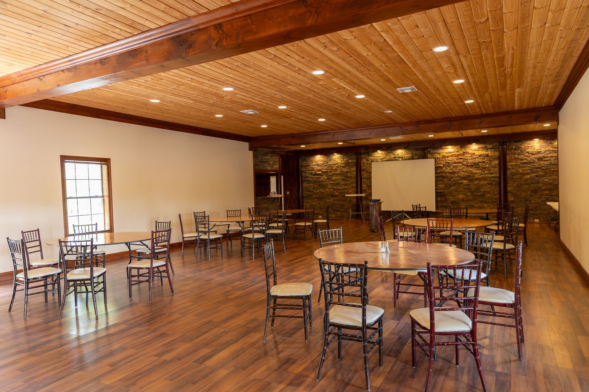 Empty event room with wood floors, tables, chairs, and a screen; wood paneled ceiling.