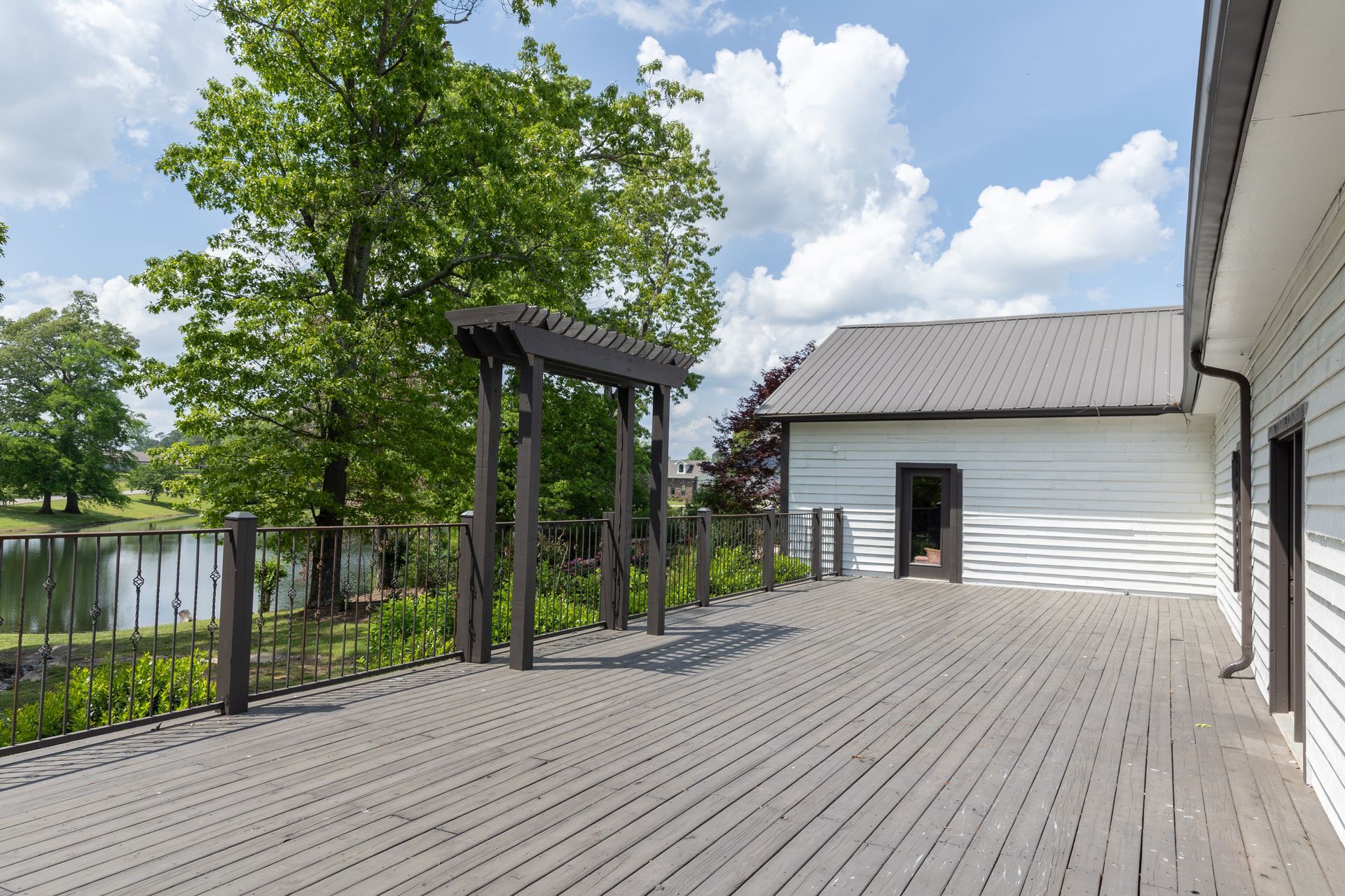 Wooden deck with a dark pergola, overlooking a lake, next to a white building.