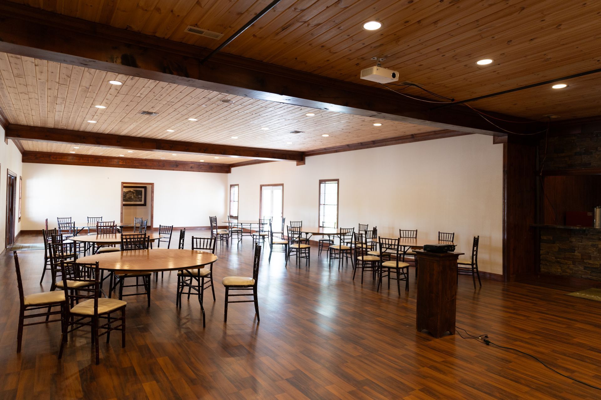 Empty event space with round tables, chairs, and wood flooring; recessed lighting, wood beam ceiling.