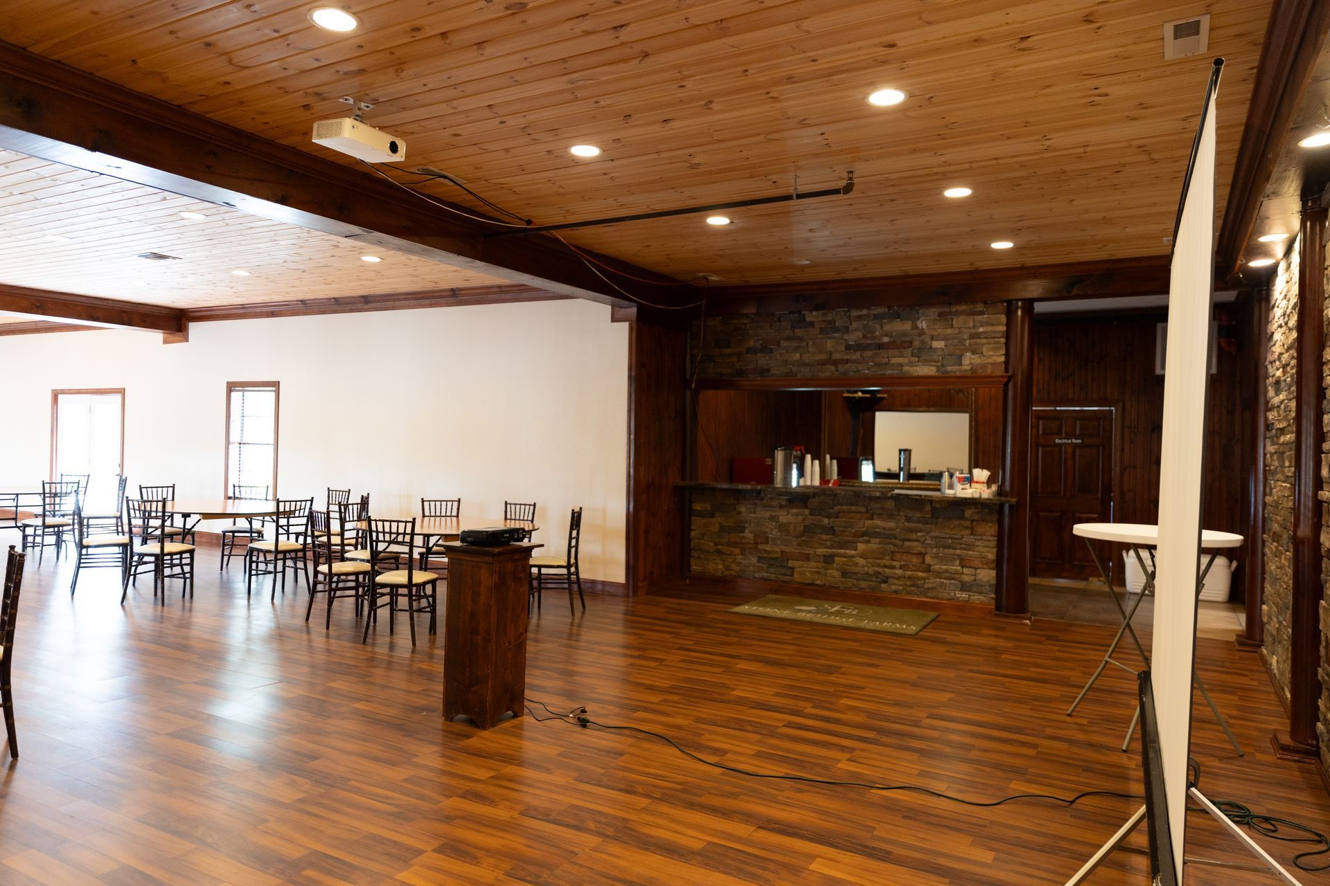 A spacious room with tables, a bar, and projection screen. Hardwood floors, wood panel ceiling, and recessed lighting.