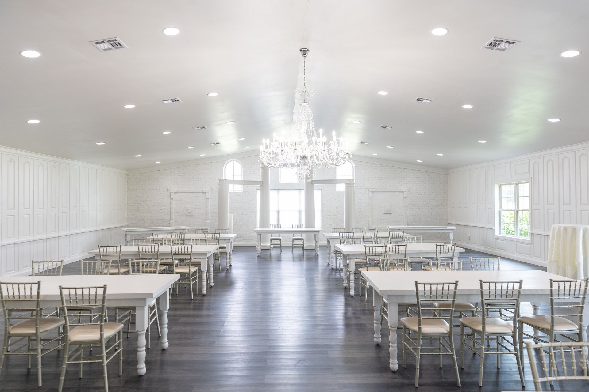 Empty white event space with tables, chairs, and chandelier. Dark wood floor, bright lighting.