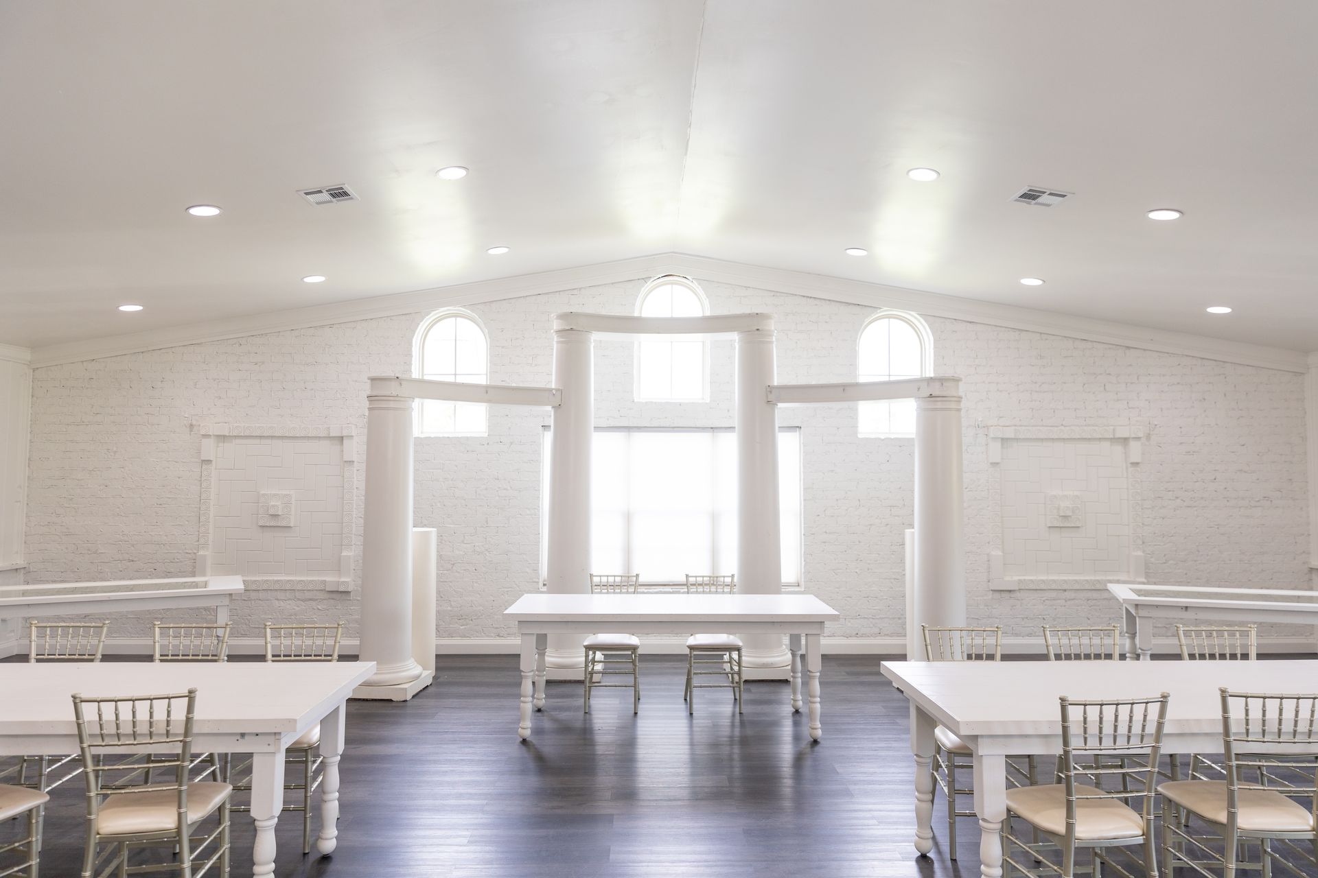 White, bright event space with tables, chairs, dark floor, and arched windows.