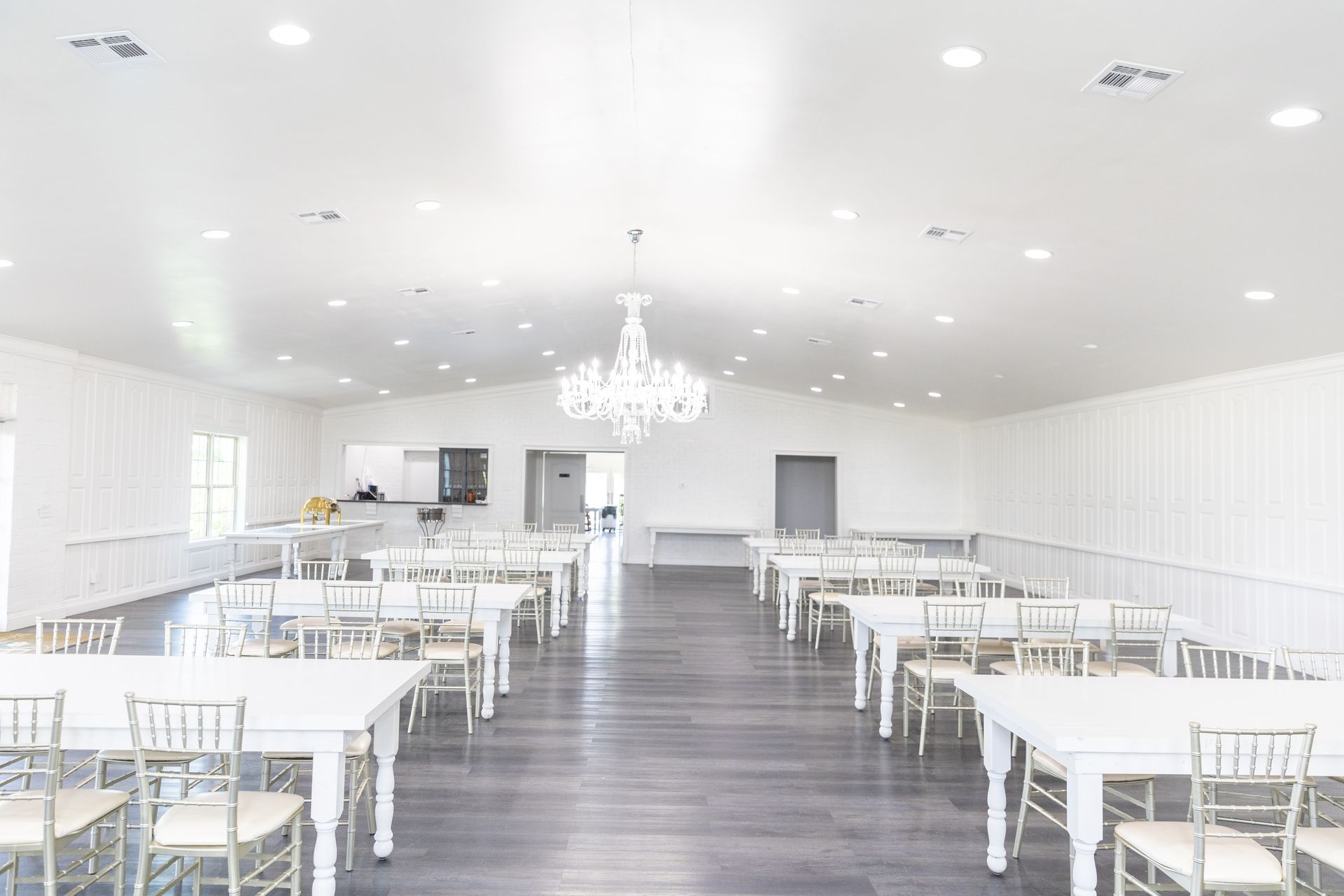 Empty event hall with white tables and chairs, wood floor, and a chandelier.