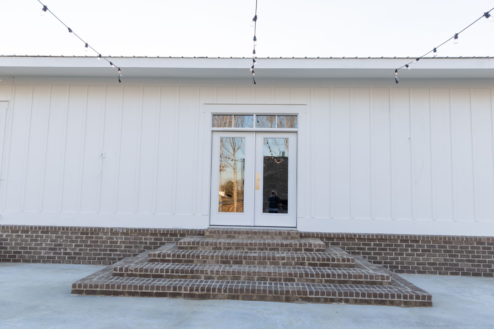 White building with brick steps leading to glass double doors. String lights above.