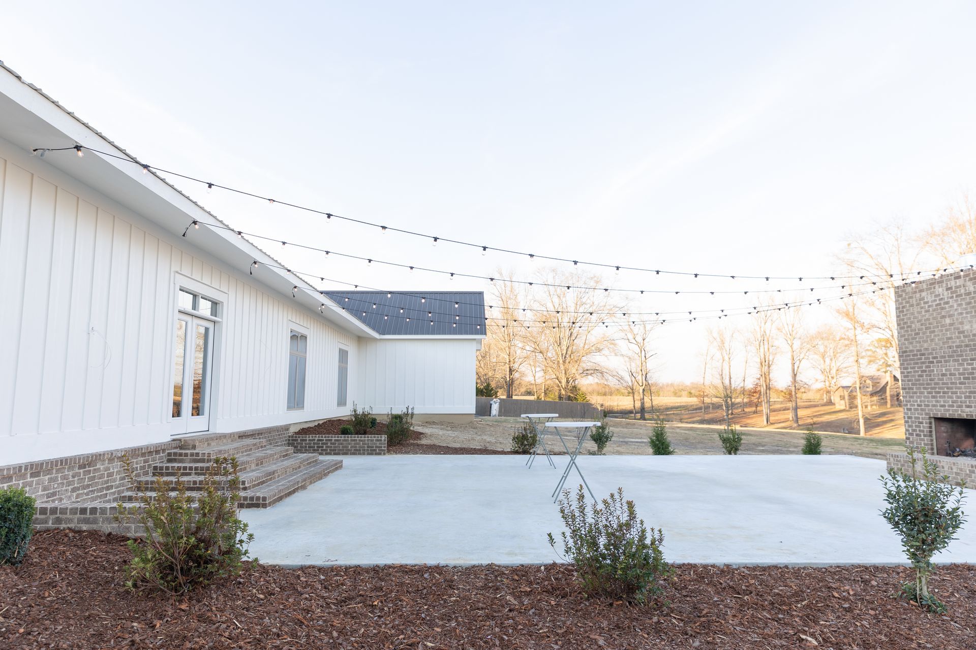 White building with concrete patio, string lights, and brick fireplace in outdoor setting.