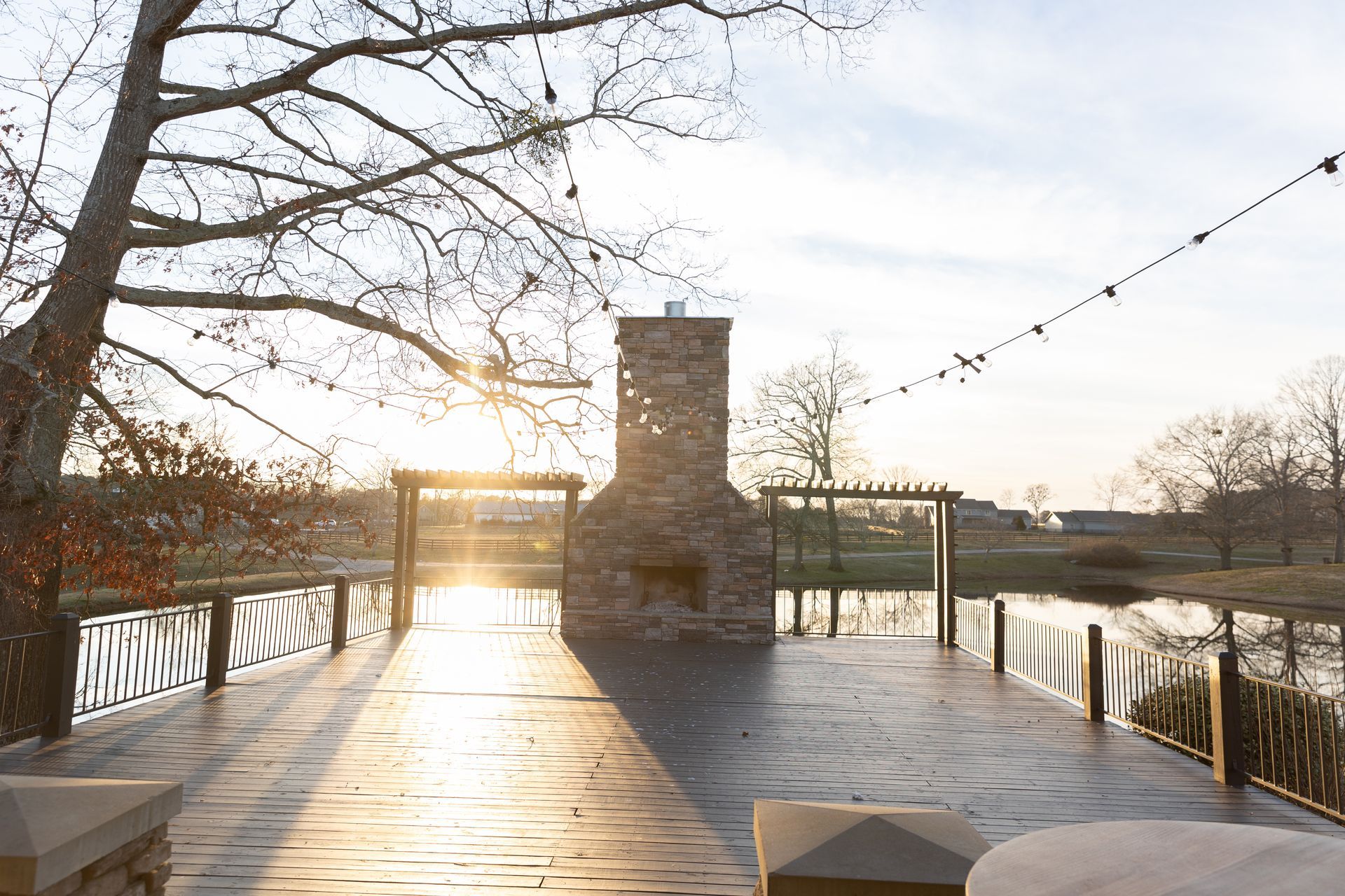 Outdoor patio with fireplace, overlooking a pond at sunset. String lights and bare trees.