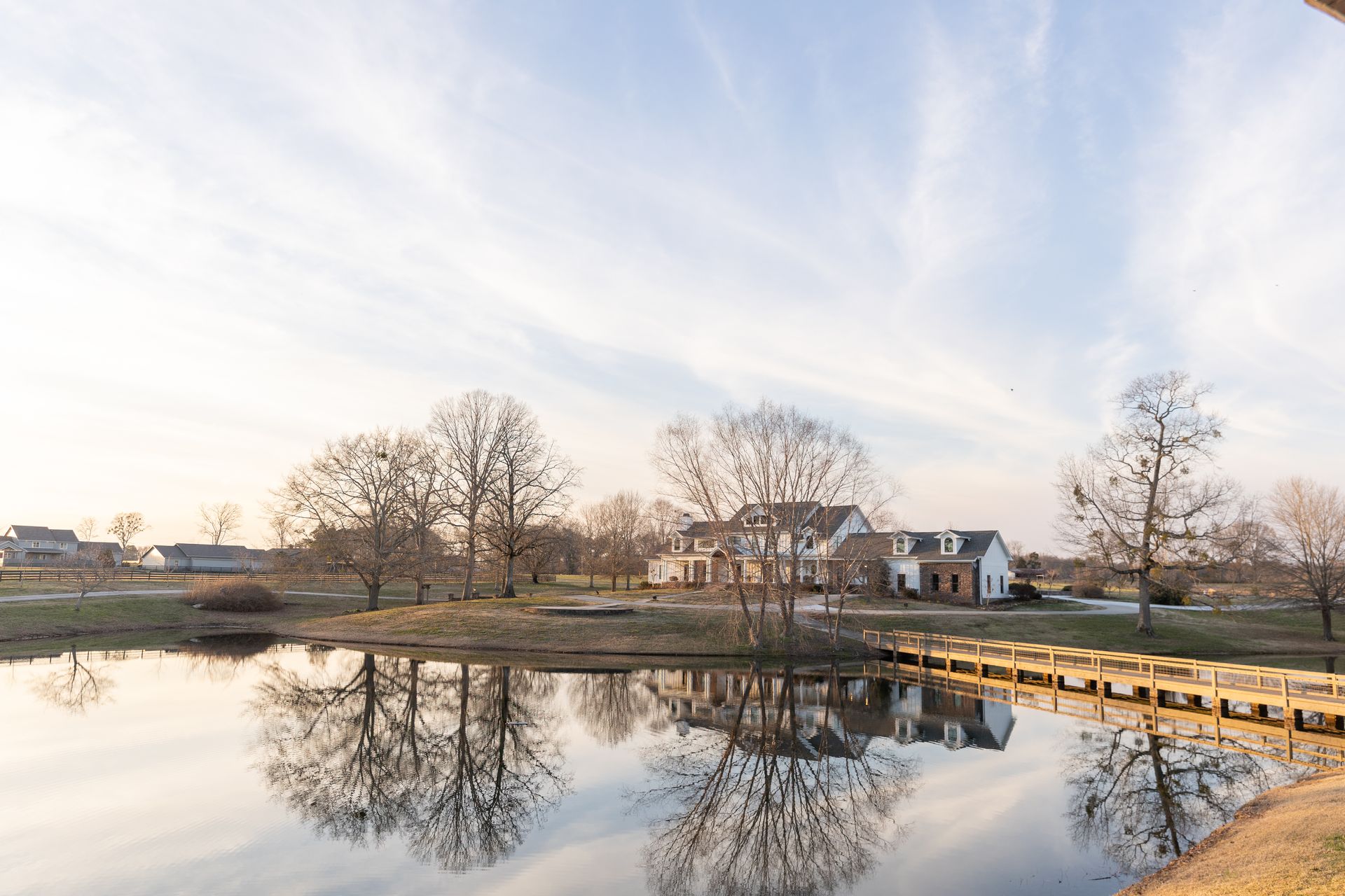 Pond with bridge reflects trees and a white house under a cloudy sky.